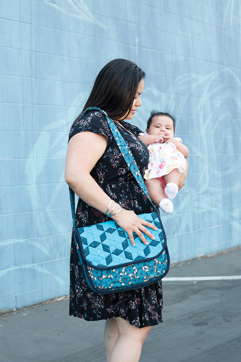 A woman in a black floral dress holds a baby outdoors against a blue mural, carrying Stash Books’ Fabric Weaving shoulder bag, its blue quilted geometric pattern showcasing fine handcrafted woven textiles.