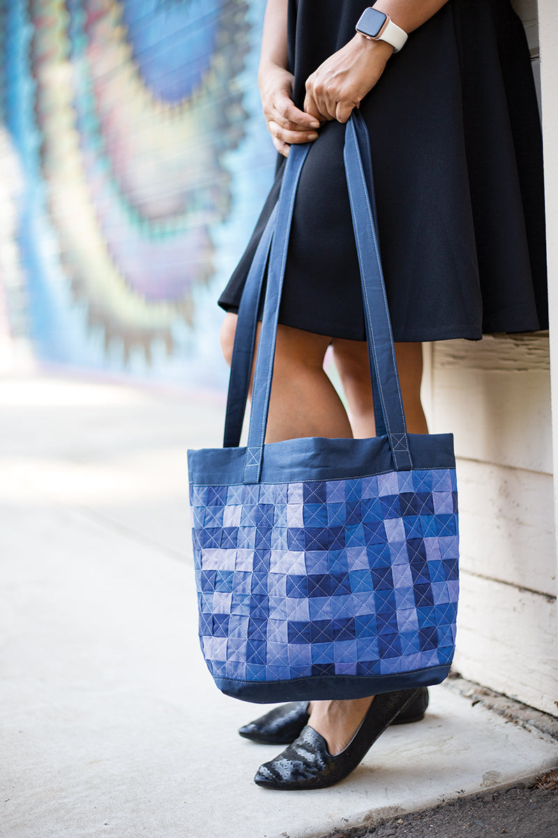 A person in a black dress and flats holds the "Fabric Weaving" tote bag by Stash Books, featuring handcrafted geometric patterns, while standing on a sidewalk beside a colorful mural.
