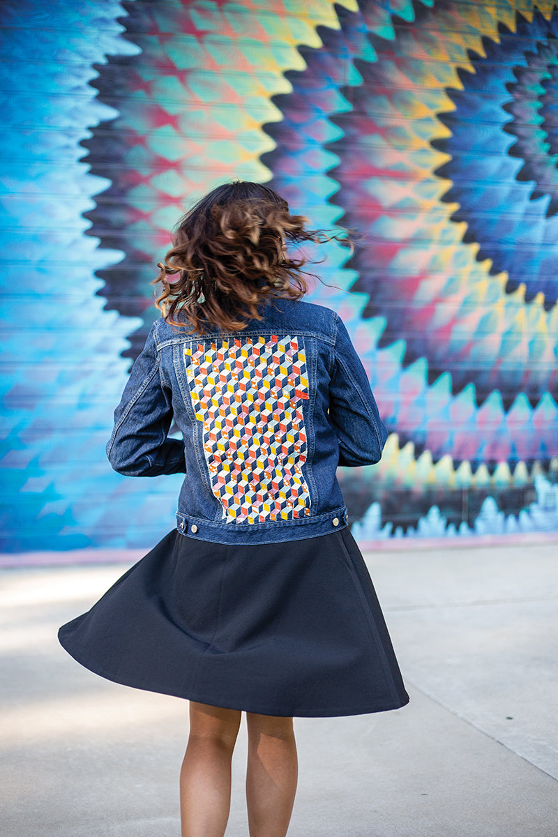 In front of a vibrant mural with blue, yellow, and red spirals, a woman in a black skirt twirls while wearing a blue denim jacket featuring a geometric patch inspired by Fabric Weaving from Stash Books.
