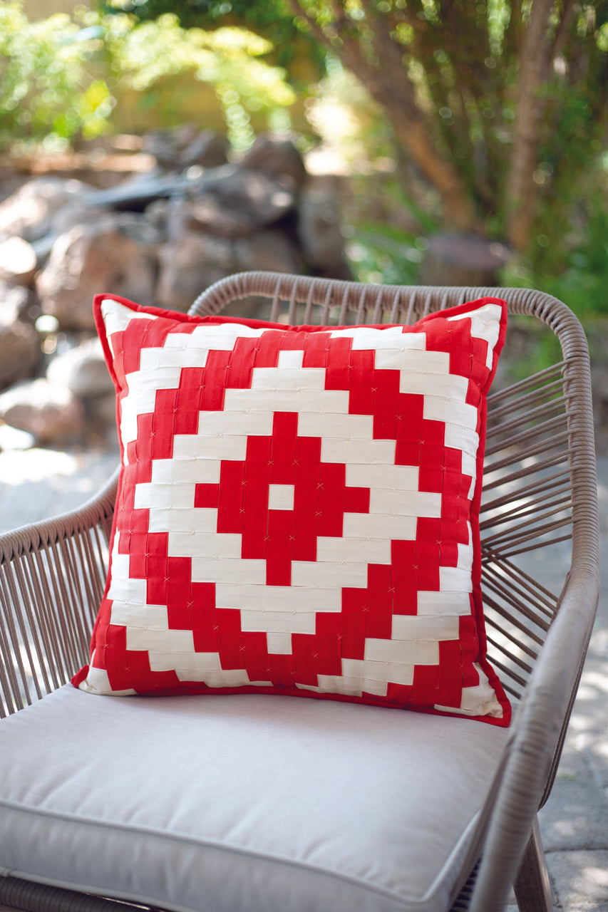 A Fabric Weaving cushion by Stash Books, featuring a bold red and white geometric design, sits on a light gray outdoor chair with plants and rocks in the sunlit background.