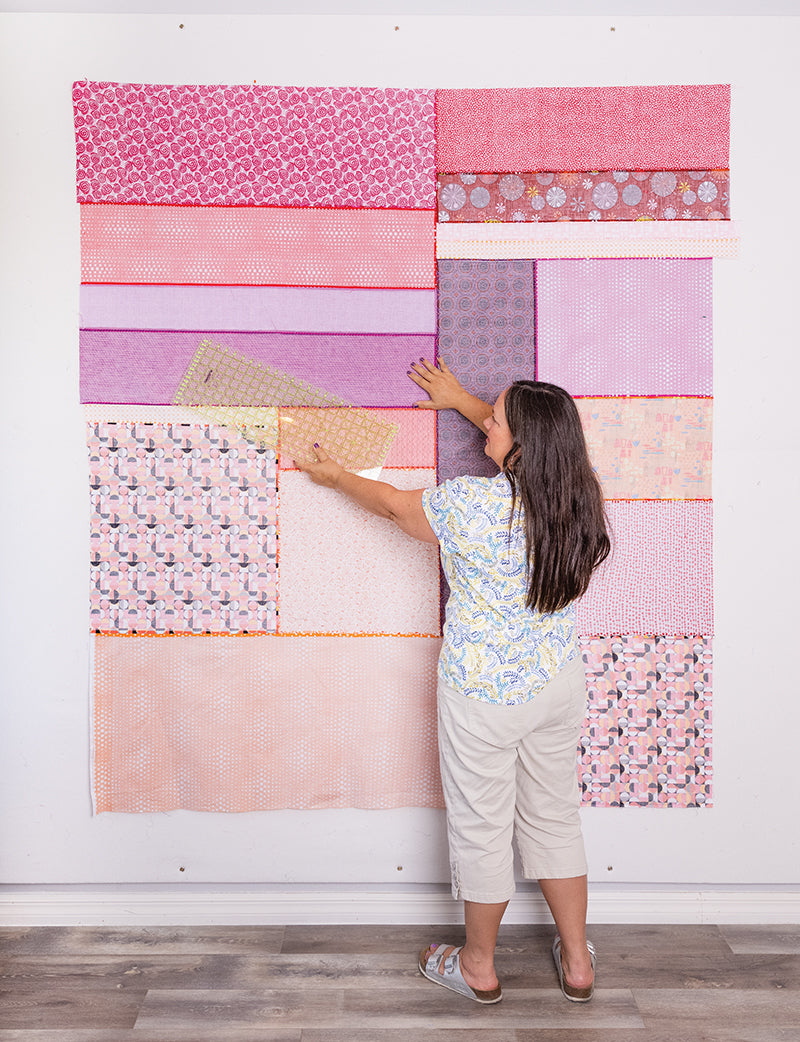 A woman arranges pastel rectangles on a white wall, creating a modern patchwork design inspired by "How Do I Quilt It?" from Stash Books. She stands barefoot on a wooden floor, facing the quilt pieces in pink, purple, and peach tones.
