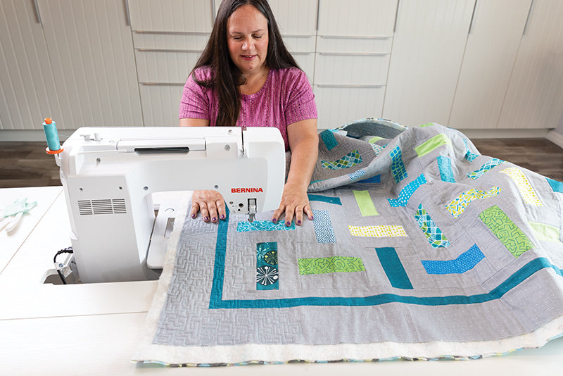 A woman with long brown hair wearing a pink shirt is quilting a colorful patchwork using free-motion techniques from "How Do I Quilt It?" by Stash Books on a white sewing machine in a bright, modern sewing room.