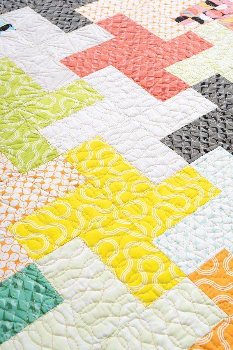 Close-up of the "How Do I Quilt It?" quilt by Stash Books, showcasing plus-shaped patterns in yellow, green, white, peach, and black fabrics with geometric and swirly designs, stitched together using modern machine quilting.