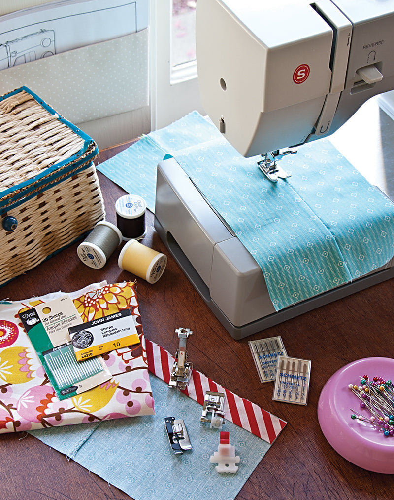 On a wooden table, a sewing machine with blue fabric is ready for a beginner project inspired by “Make Your First A-Line Skirt” from Stash Books. Colorful fabrics, spools, feet, needles, and a pink pin cushion complete the setup.