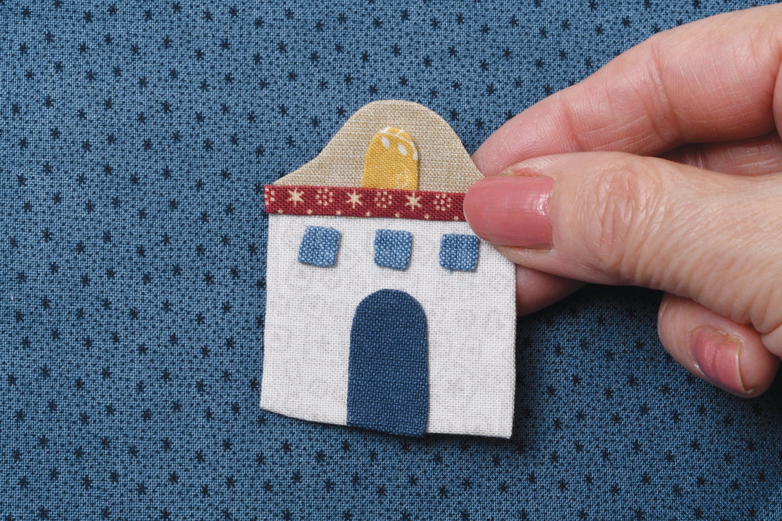 A hand holds Stash Books' Premium Hand-Appliqué Paper from Masako Wakayama, crafted into a house with a blue door, three blue windows, red roof trim, and a yellow roof arch, set against a blue dotted background.