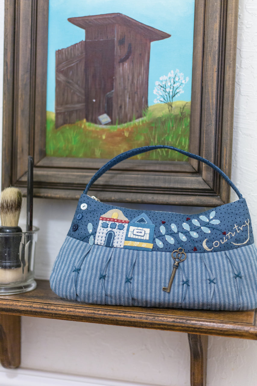 A sheet of Premium Hand-Appliqué Paper from Masako Wakayama by Stash Books sits on a wooden shelf, next to a vintage shaving brush and cup, with a framed painting of an old wooden outhouse in the background.