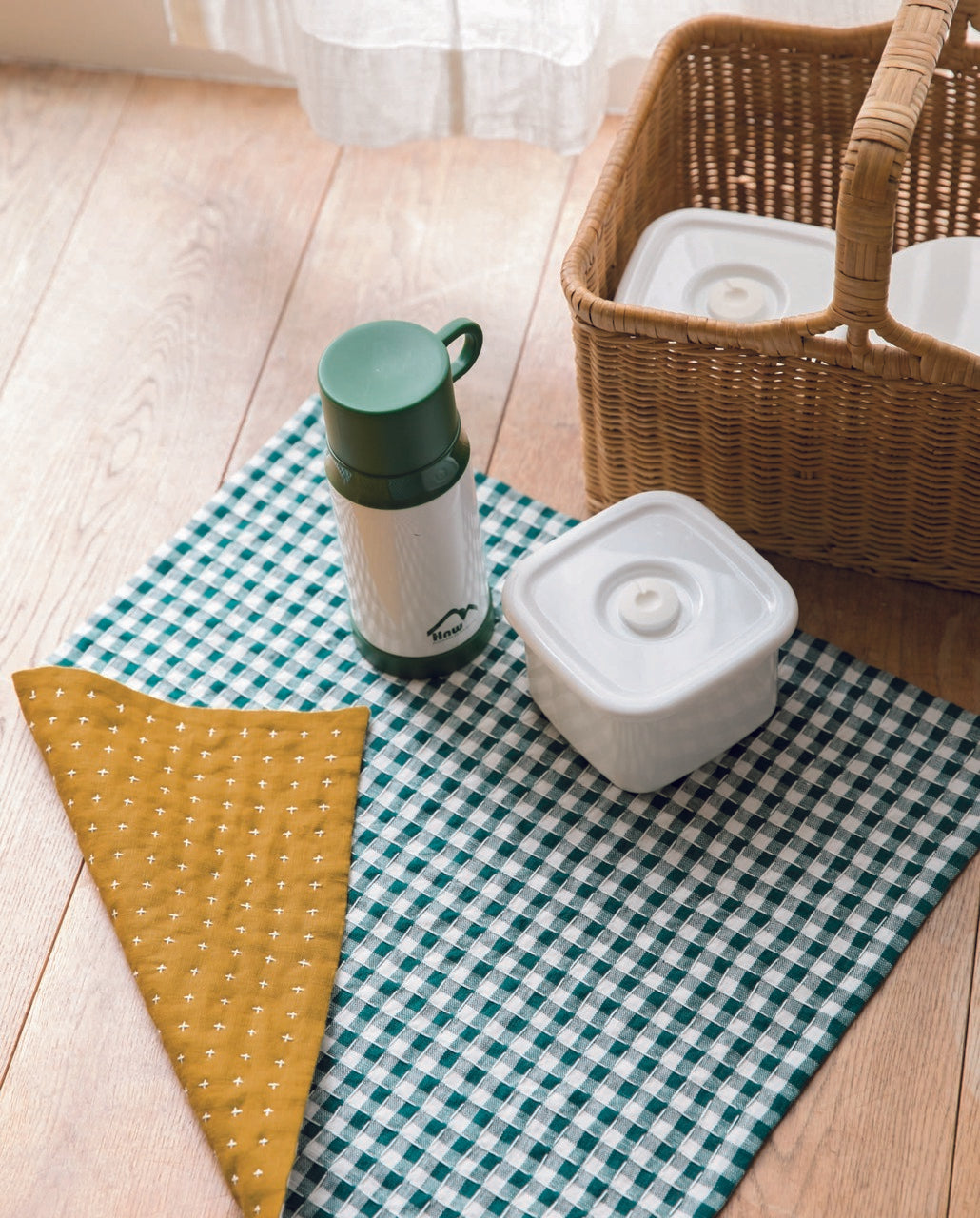 A picnic setup with Sashiko + Color by Stash Books: a green checkered cloth with subtle Sashiko stitching, a wicker basket, two white food containers, a white thermos with green lid, and a yellow cloth on a wooden floor by sheer-curtained windows.