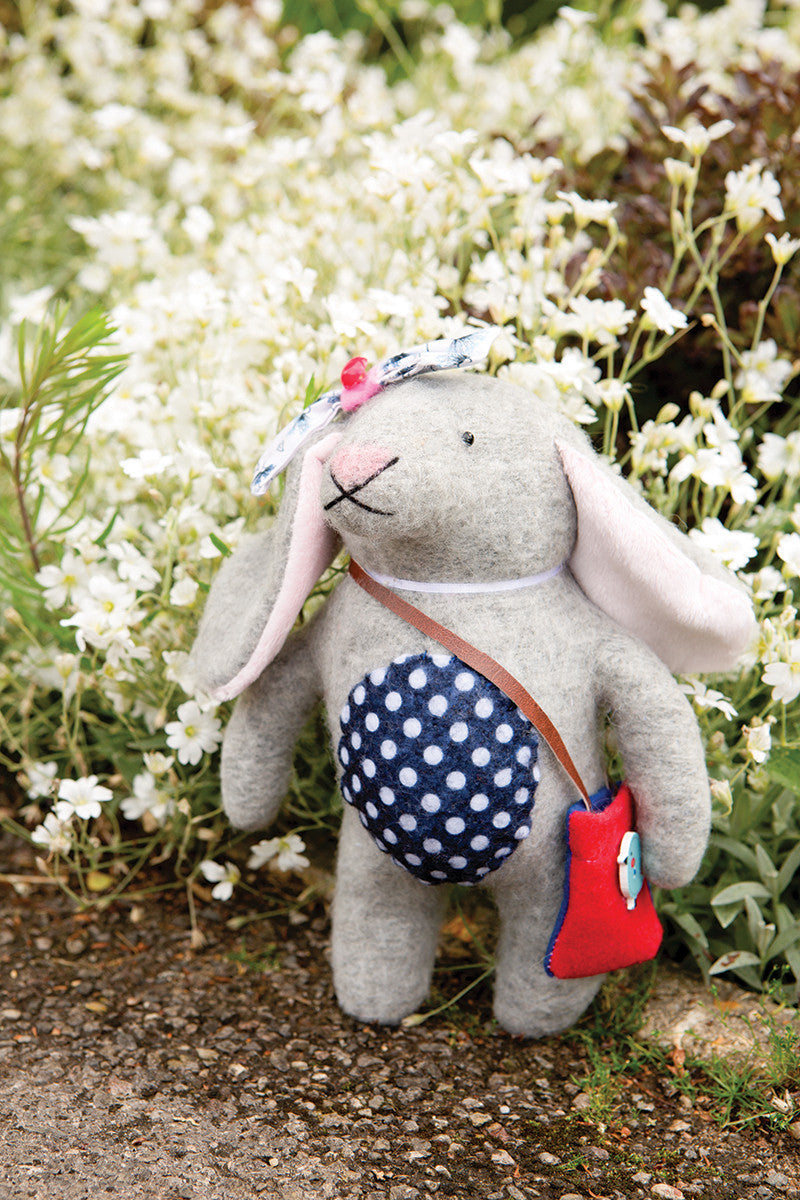 The "Instant Softies" stuffed bunny from Stash Books, featuring a blue polka-dot belly, floral headband, and red bag, stands amid white flowers—ideal inspiration for beginners looking to create their own soft toys.