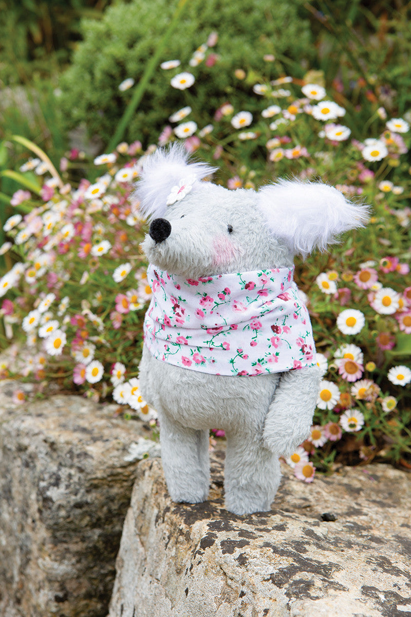 The Instant Softies plush gray koala from Stash Books, featuring pink cheeks and a floral scarf, stands among daisies—ideal inspiration for beginner soft toy crafts or anyone looking for charming handmade stuffed animals.