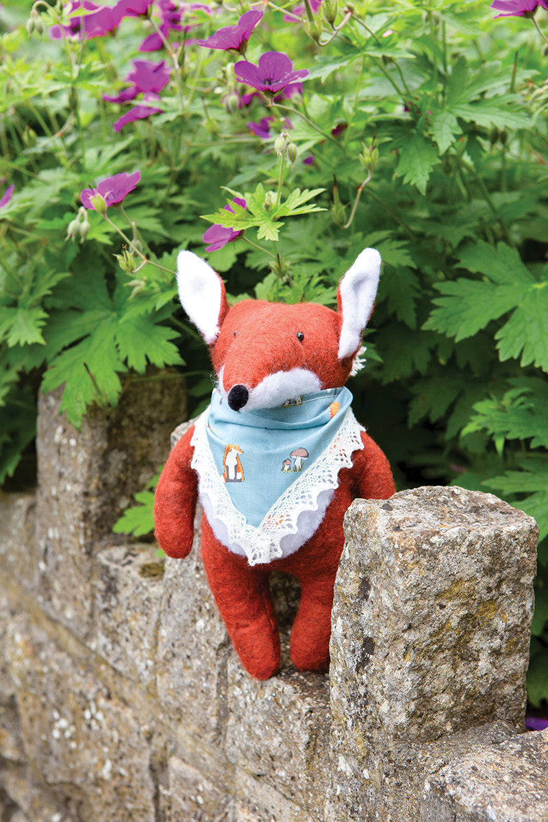 A plush fox toy with a blue handkerchief from Stash Books' "Instant Softies" rests on a stone wall among green leaves and purple flowers—a delightful handmade stuffed animal for beginner soft toy crafters.