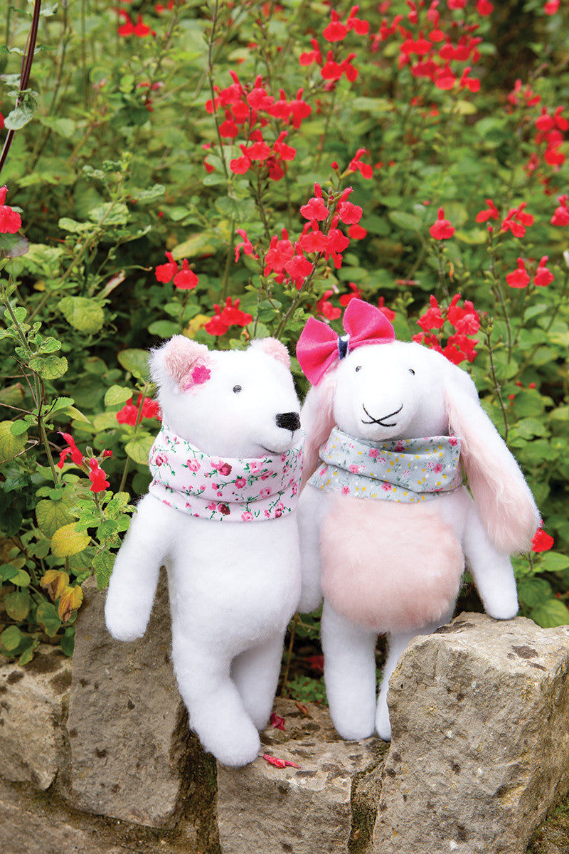 Two handmade stuffed animals from "Instant Softies" by Stash Books—a white bear and a pink-eared rabbit with patterned scarves—stand side by side among red flowers, inspiring beginner soft toy crafters.
