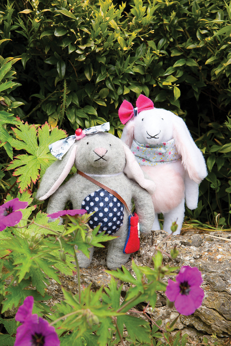 Two Instant Softies plush bunny toys by Stash Books stand outdoors among plants and purple flowers. One gray bunny has a headband and blue polka-dot bag; the white bunny wears a pink scarf and bow, surrounded by lush foliage.