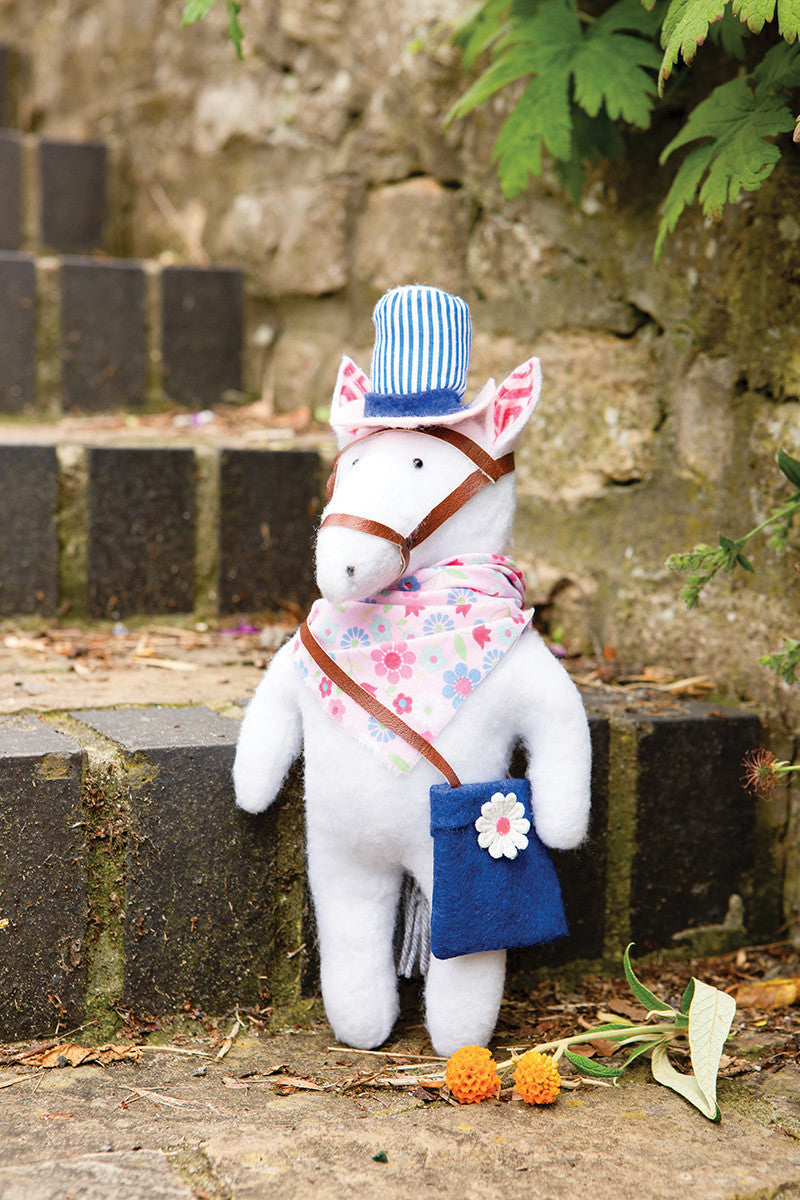 Instant Softies by Stash Books features a handmade white horse plushie with a blue-striped hat, floral scarf, and blue flower bag. Shown on stone steps with greenery and orange pom-poms—great inspiration for beginner soft toy creators.