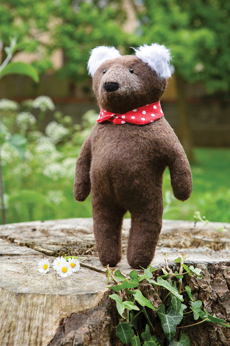 A handmade brown teddy bear from Stash Books' Instant Softies stands upright on a tree stump outdoors, wearing a red polka dot scarf, with daisies and green ivy nearby and a blurred garden in the background.