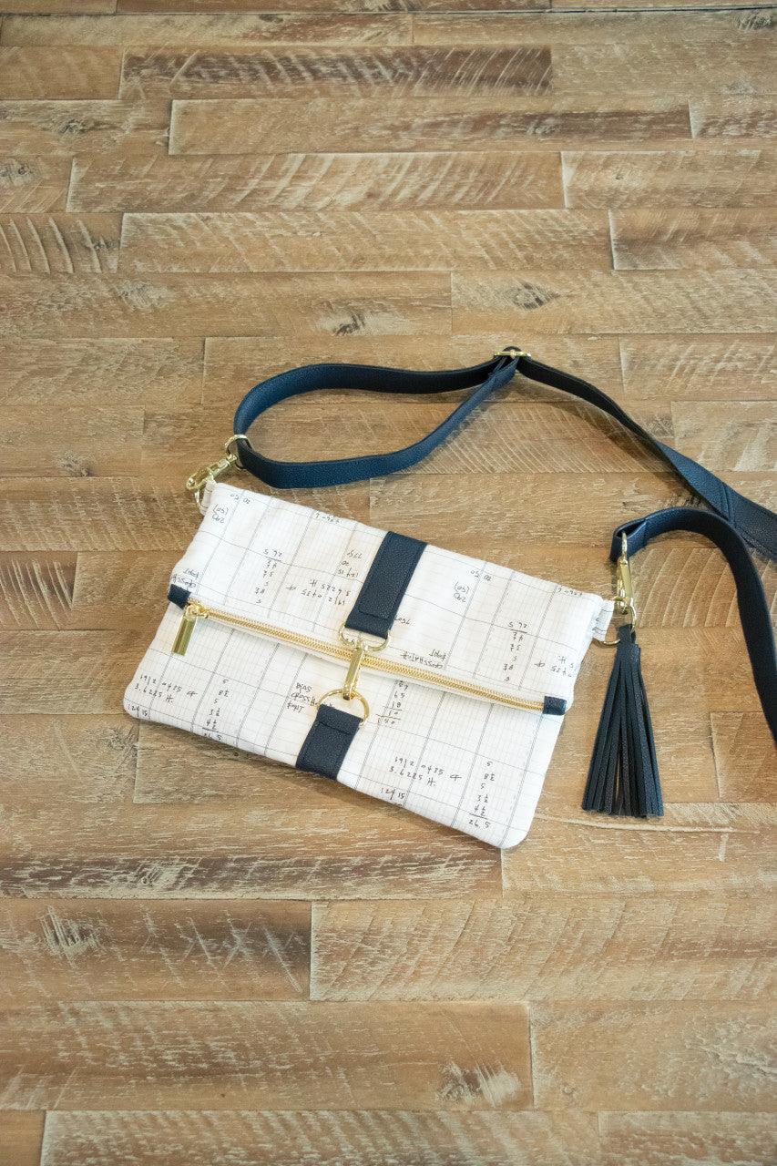 Making Bags, A Field Guide by C&T Publishing rests on a wooden floor, its beige crossbody design featuring black straps, gold hardware, a zipper, and a black tassel with expert sewing techniques for professional results.