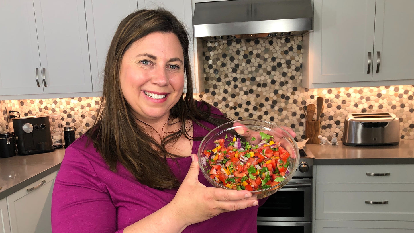 A woman in a purple top smiles in a modern kitchen with white cabinets, holding a bowl of freshly made salsa to showcase her Next Level Nachos course by Thinkific. A patterned backsplash completes the inviting scene.
