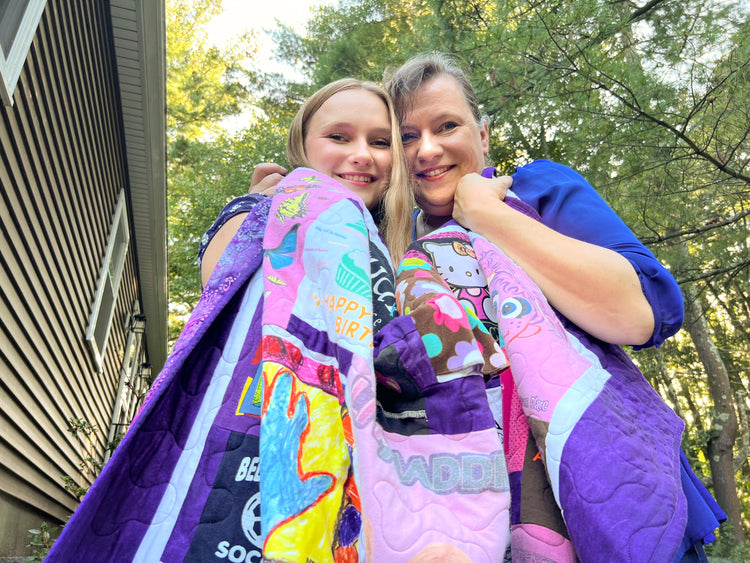 A smiling woman and girl stand close together outside, wrapped in colorful, patterned quilts, with trees and a house in the background.