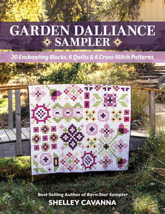 The Garden Dalliance Sampler by C&T Publishing, featuring vibrant pink, purple, green, and white geometric patterns, is draped over a wooden bridge in a sunlit garden—perfect inspiration for your next quilting project.