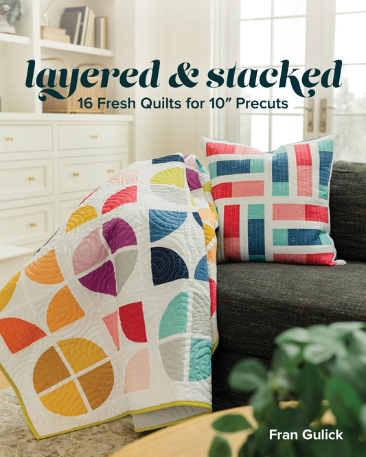 A vibrant quilt and matching pillow made with 10″ precuts adorn a dark sofa. Text: Layered & Stacked by Stash Books—16 fresh quilts for 10″ precuts by Fran Gulick.