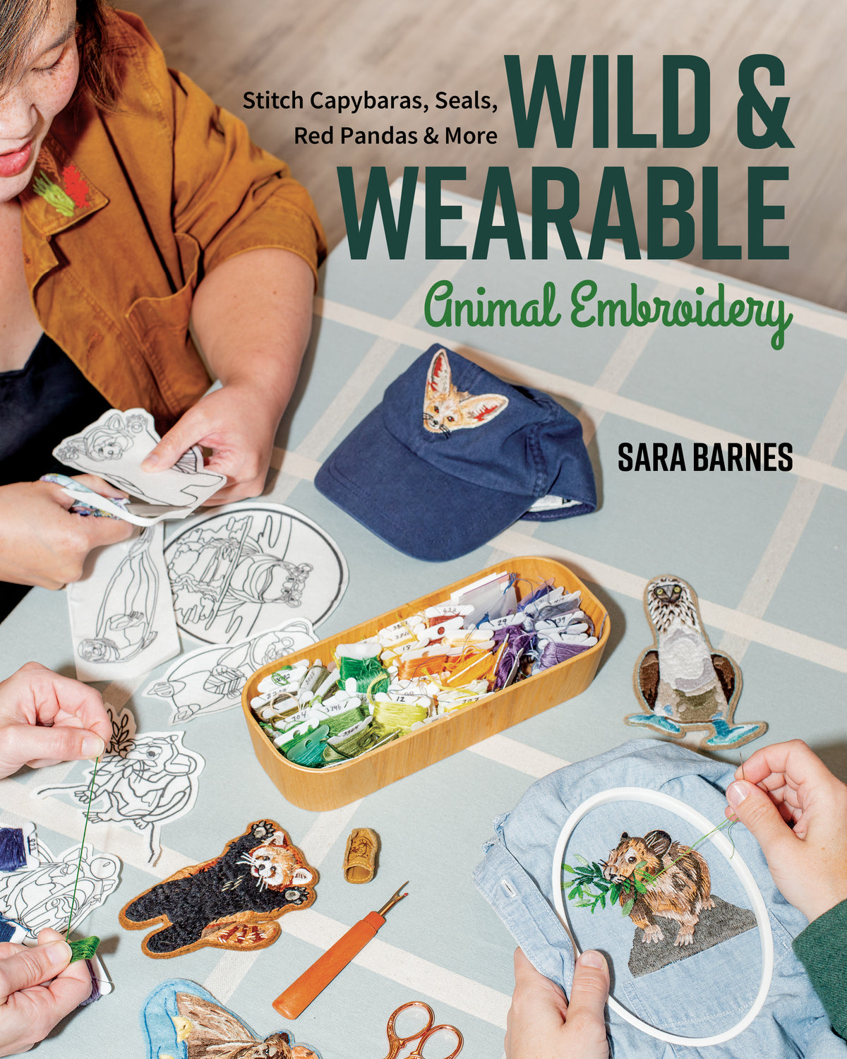 Hands stitch and prepare Wild & Wearable Animal Embroidery patches on a table filled with thread, hoops, and a baseball cap to create sustainable wardrobe accessories.