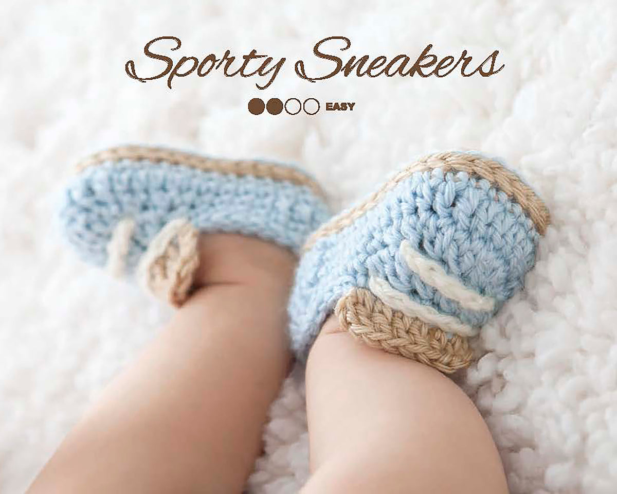 A close-up of a baby’s feet in blue and beige crocheted booties on a soft white blanket, with “Modern Booties for Baby” by Leisure Arts—an easy beginner crochet project perfect for fans of their designs.
