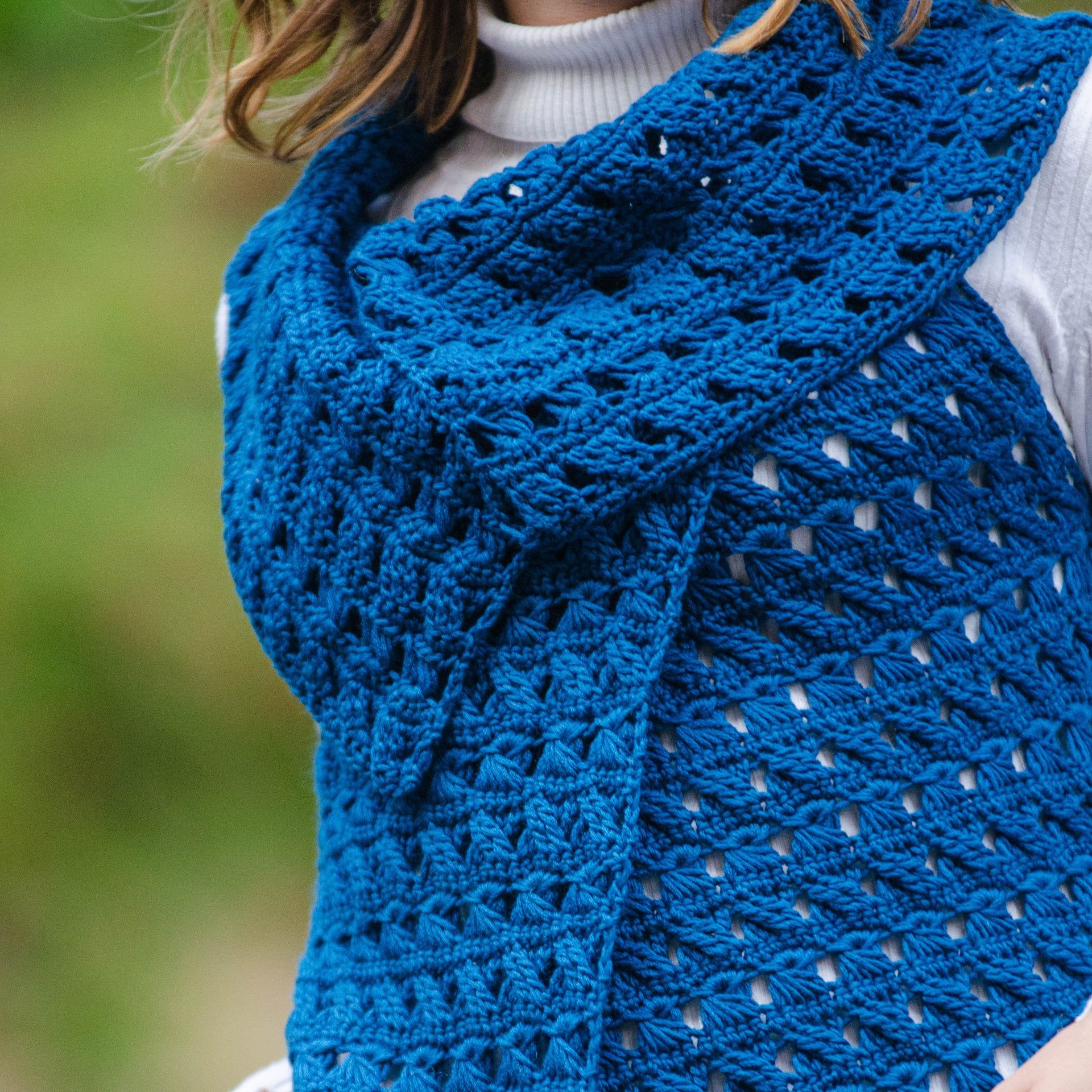 A person wears Creative Sparks Non-Stick Crochet Lace shawl, featuring an intricate blue pattern over a white turtleneck. The elegant drape and textured stitches complement the blurred greenery backdrop.