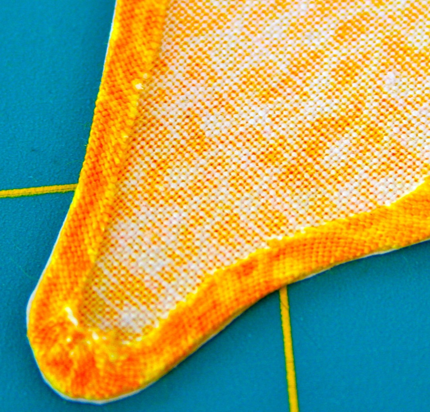 A close-up of the Innovative Appliqué by Creative Spark displays a textured, triangular fabric piece with vibrant orange edges set against a teal grid background, highlighting intricate details reminiscent of stunning quilts.