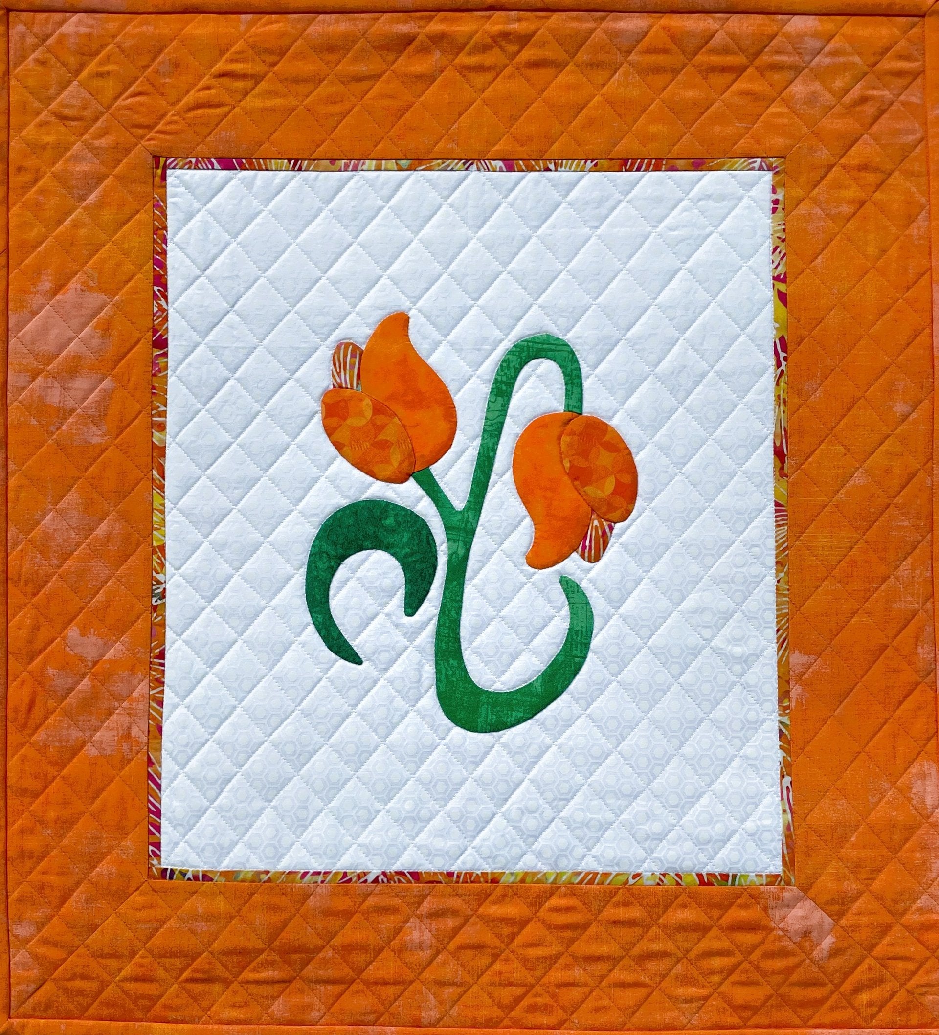 The Innovative Appliqué quilt by Creative Spark features a stunning orange design with a white center panel adorned with two orange flowers and green stems. It includes a grid texture, surrounded by an elegant solid orange border with a quilted diamond pattern.