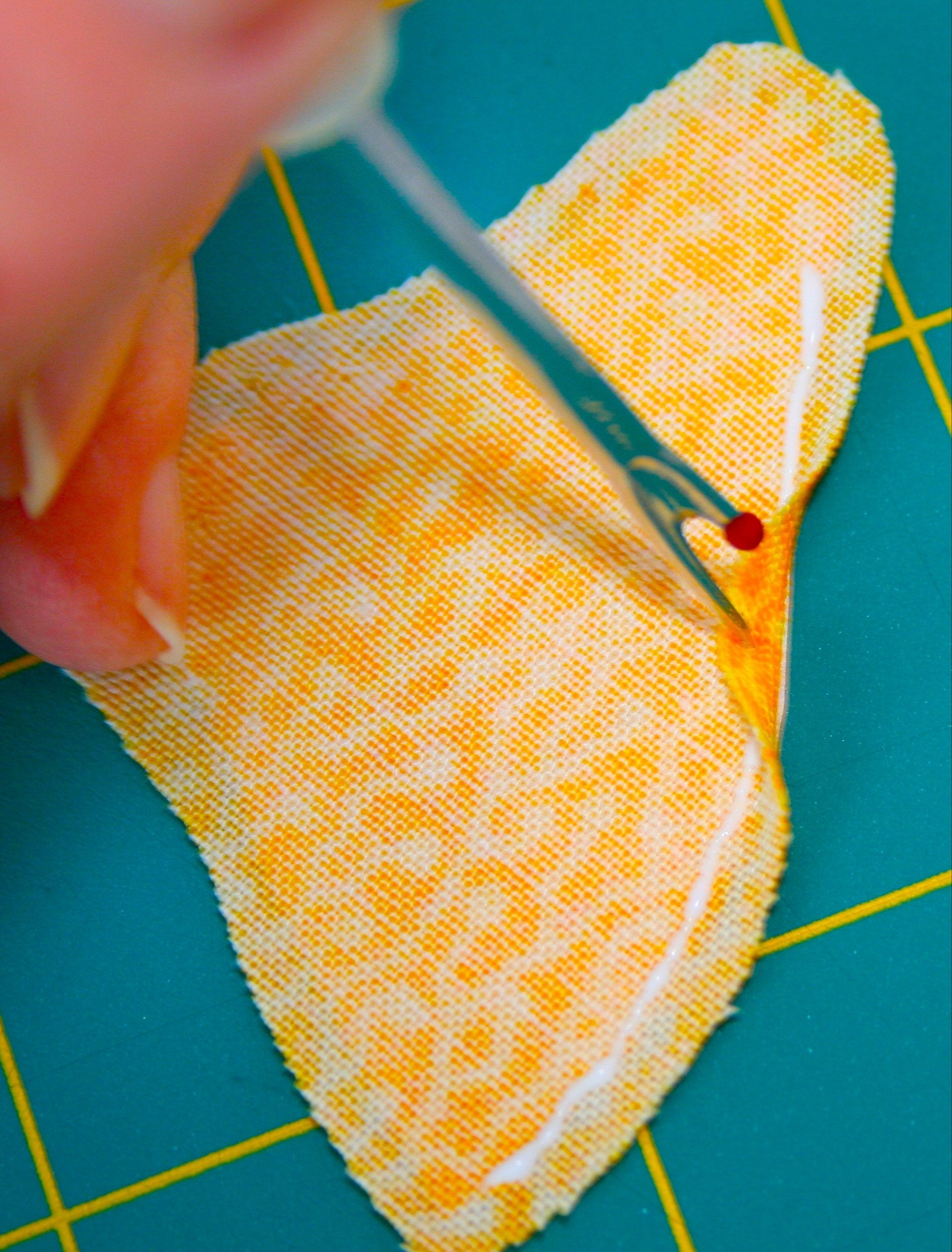 A person uses the Innovative Appliqué by Creative Spark to expertly cut through bright yellow patterned fabric on a green mat, positioning the intricately detailed tool under a stitch to gently separate threads and prepare for quilts and turned-edge appliqué projects.