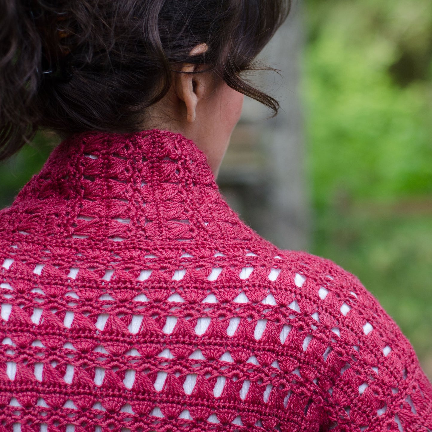 A woman with dark hair faces away from the camera, draped in a Creative Spark Non-Stick Crochet Lace shawl in vibrant pink. The intricate openwork pattern stands out against a blurred green foliage background.