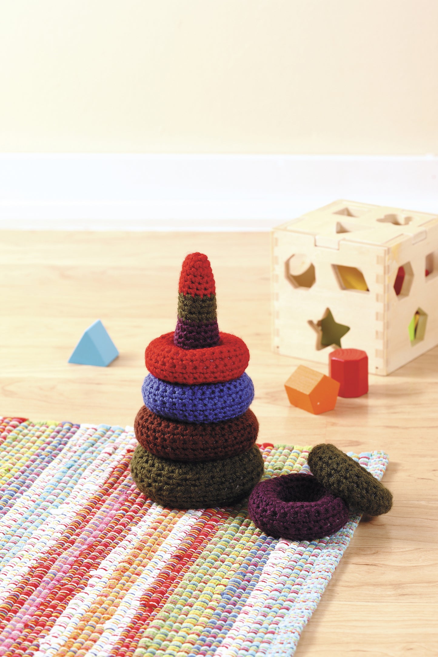 The "Everyday Crochet for All" stacking ring toy by Leisure Arts features jewel tones and easy crochet patterns. It rests on a colorful rug next to a wooden shape-sorter cube and scattered shapes on a light wood floor.