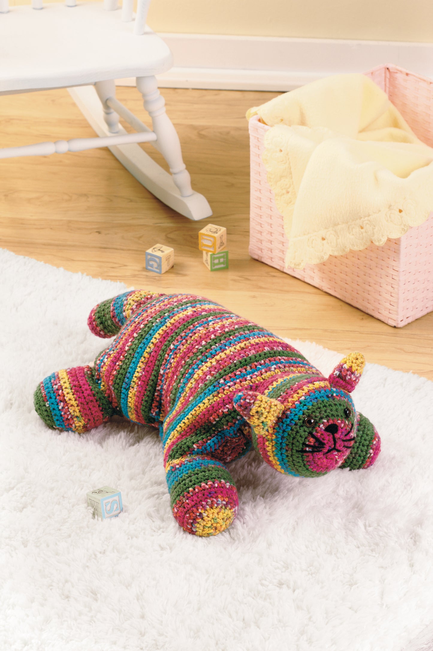 A cheerful crocheted bear from the Leisure Arts book "Everyday Crochet for All" rests on a nursery rug beside wooden letter blocks and a pink basket with a yellow blanket near the white rocking chair. Perfect for beginner projects!.