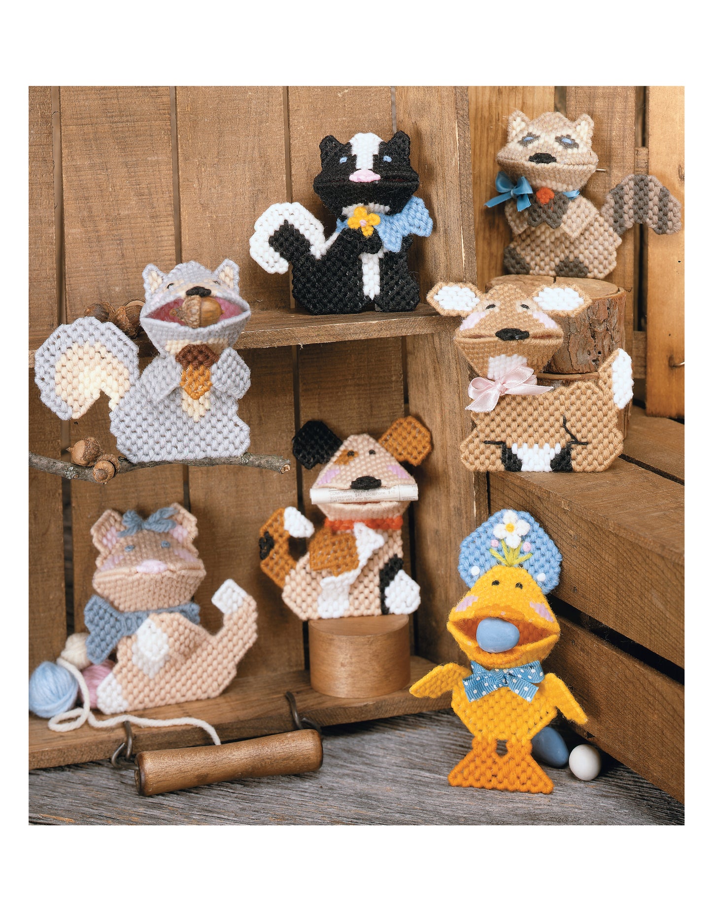 Leisure Arts’ Plastic Canvas Kissers Digital Download features handmade animal figures—raccoon, skunk, two dogs, duck, and two cats—crafted from plastic canvas patterns and displayed on shelves with yarn balls and spools.