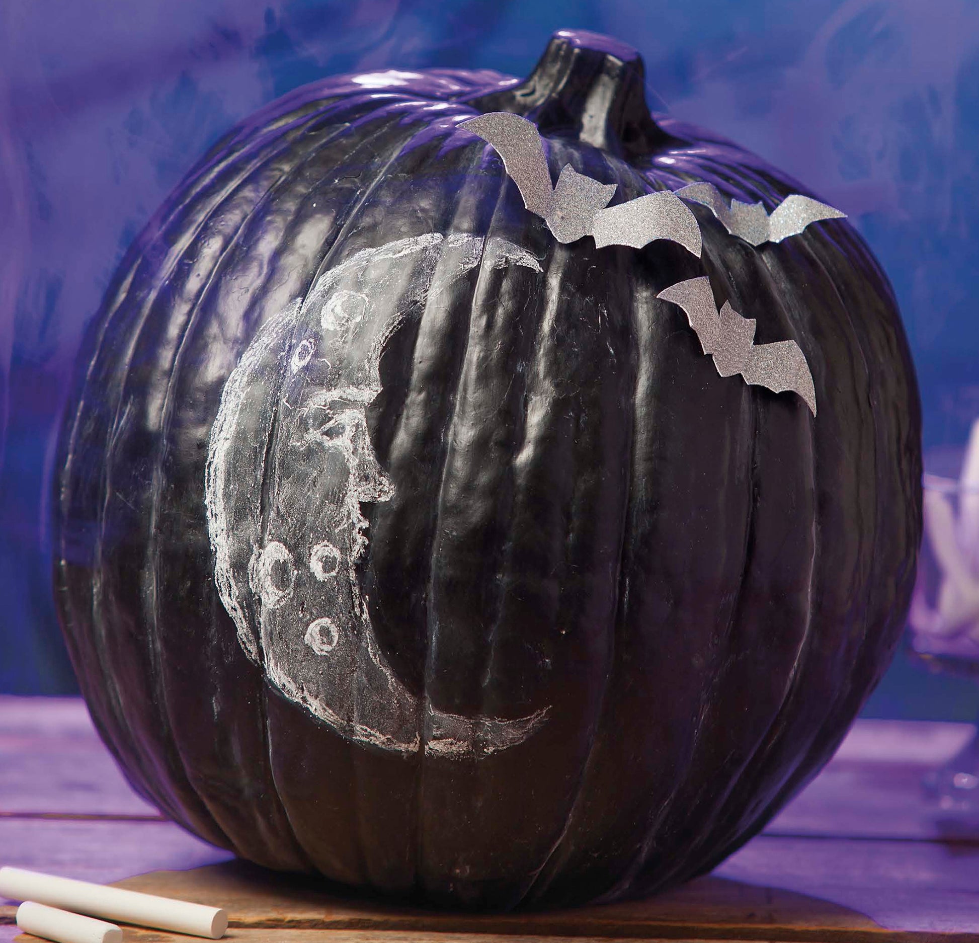 The "Pumpkins Pumpkins Pumpkins" kit by Leisure Arts includes a black faux pumpkin with a white chalk crescent moon face and craters, plus three silver paper bats—perfect for creative DIY pumpkin crafts.