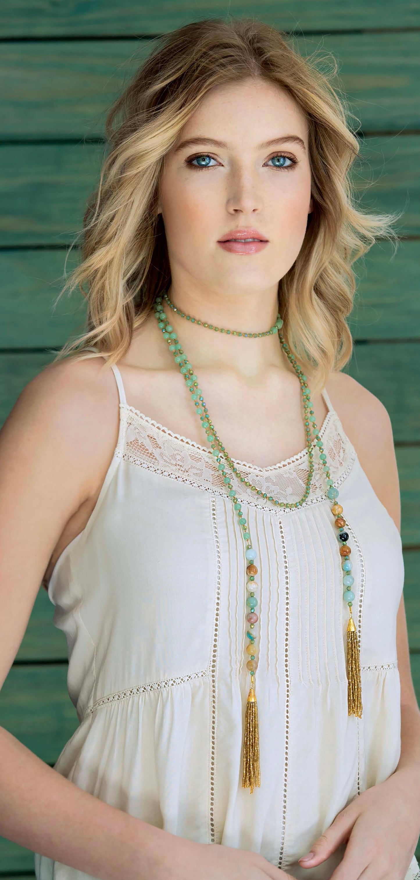 A woman with wavy blonde hair models layered green beaded fringe necklaces from Leisure Arts’ "Tasseled Jewelry—From Chains & Leather to Ribbons & Beads," wearing a sleeveless cream top in front of a green wooden background.