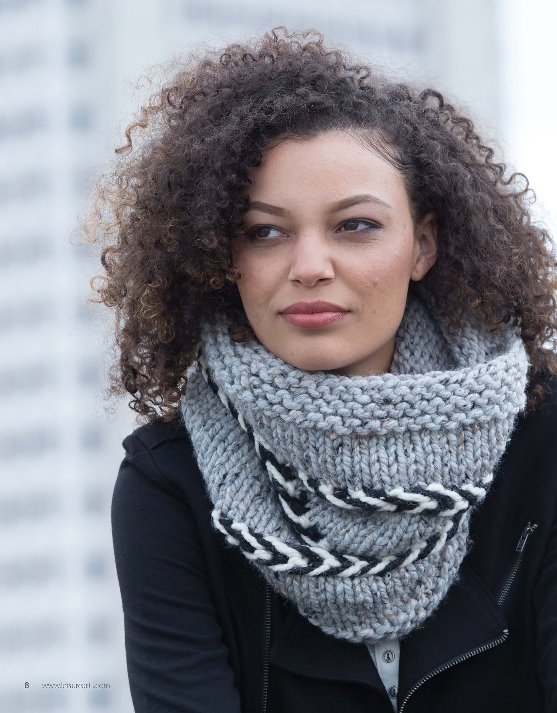 A woman with curly hair wears a gray scarf featuring black and white chevron accents, made with super bulky yarn. She's looking off to the side, with a blurred tall building behind her. Scarf pattern: "Slow Fashion Made Fast" by Leisure Arts.