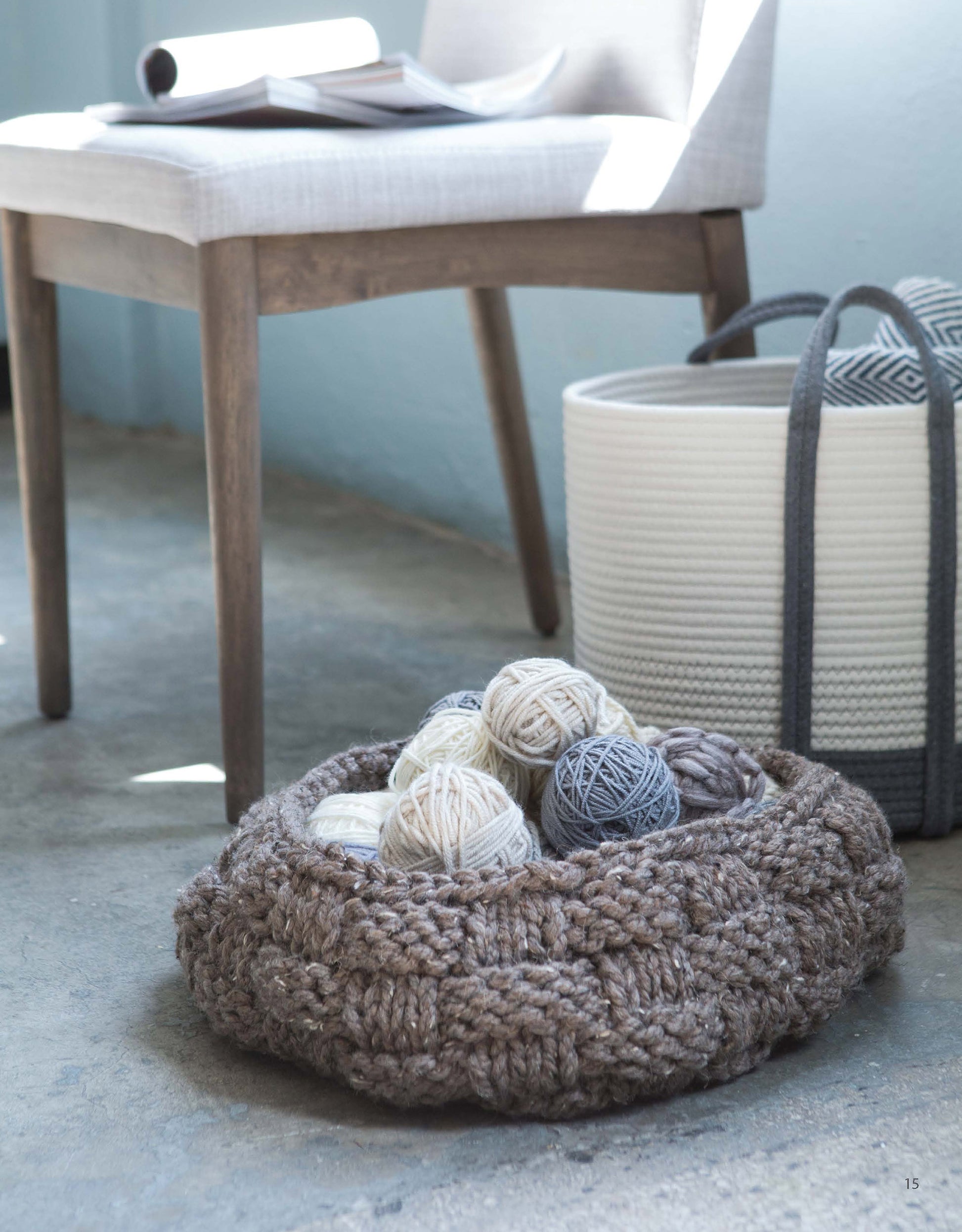 The ‘Slow Fashion Made Fast’ basket by Leisure Arts, ideal for super bulky yarn, holds cream and blue yarn balls on a concrete floor near a wooden chair. A white rope storage basket and gray tote bag appear in the background.