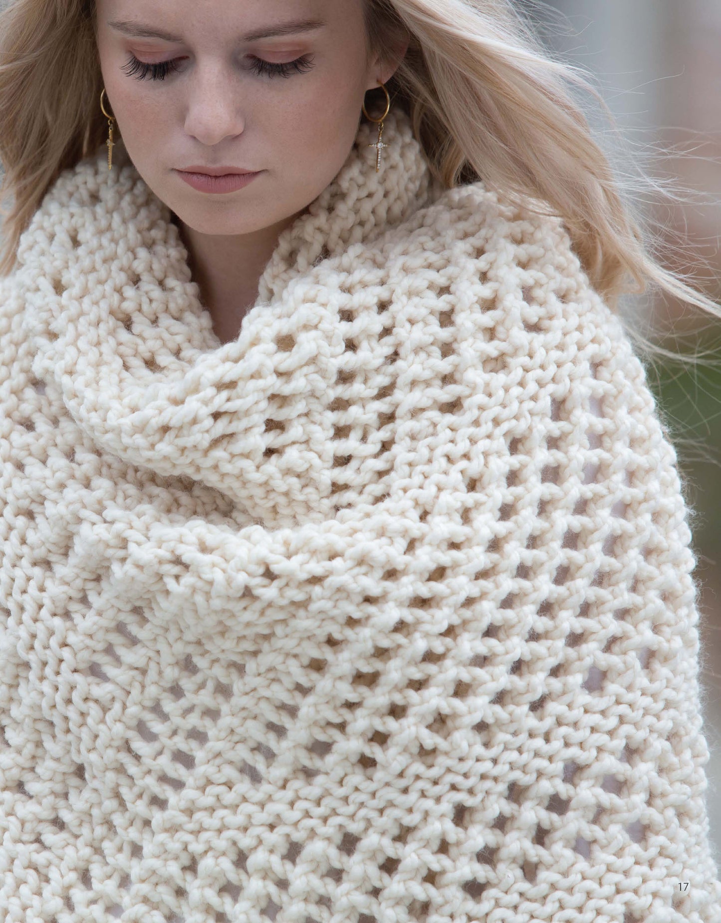 A blonde woman looks down calmly, wearing "Slow Fashion Made Fast" by Leisure Arts—a large, cream-colored shawl with a loose, chunky textured knit that drapes over her shoulders and chest.