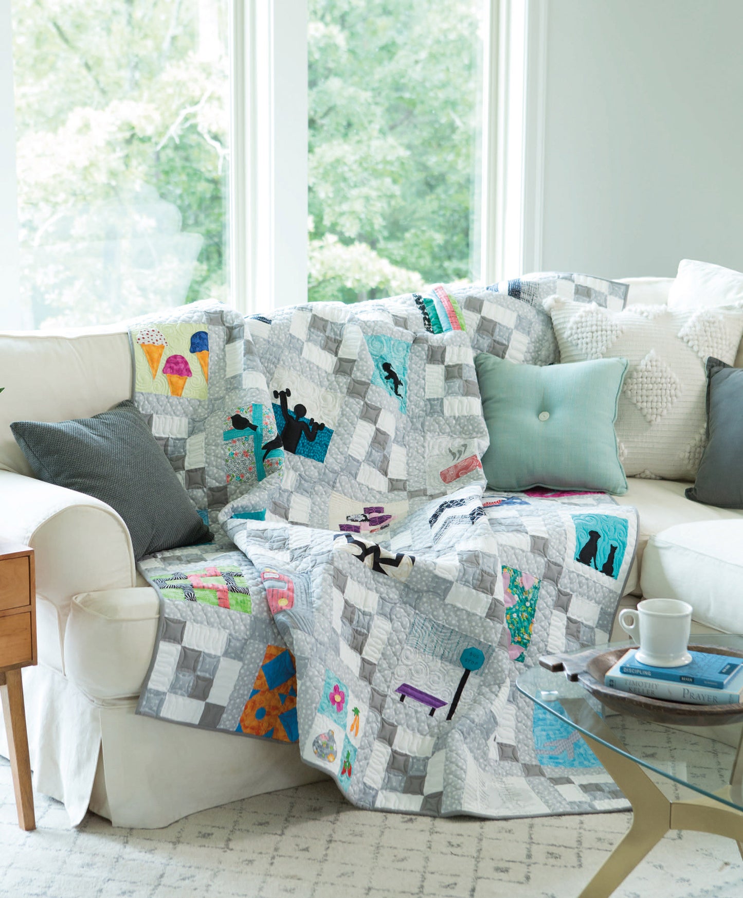 A vibrant City Blocks—32 Pieced and Applique Blocks Portraying City Life quilt from Leisure Arts is displayed over a white sectional with gray and aqua pillows in a sunlit living room, beside a side table with a coffee cup and book.