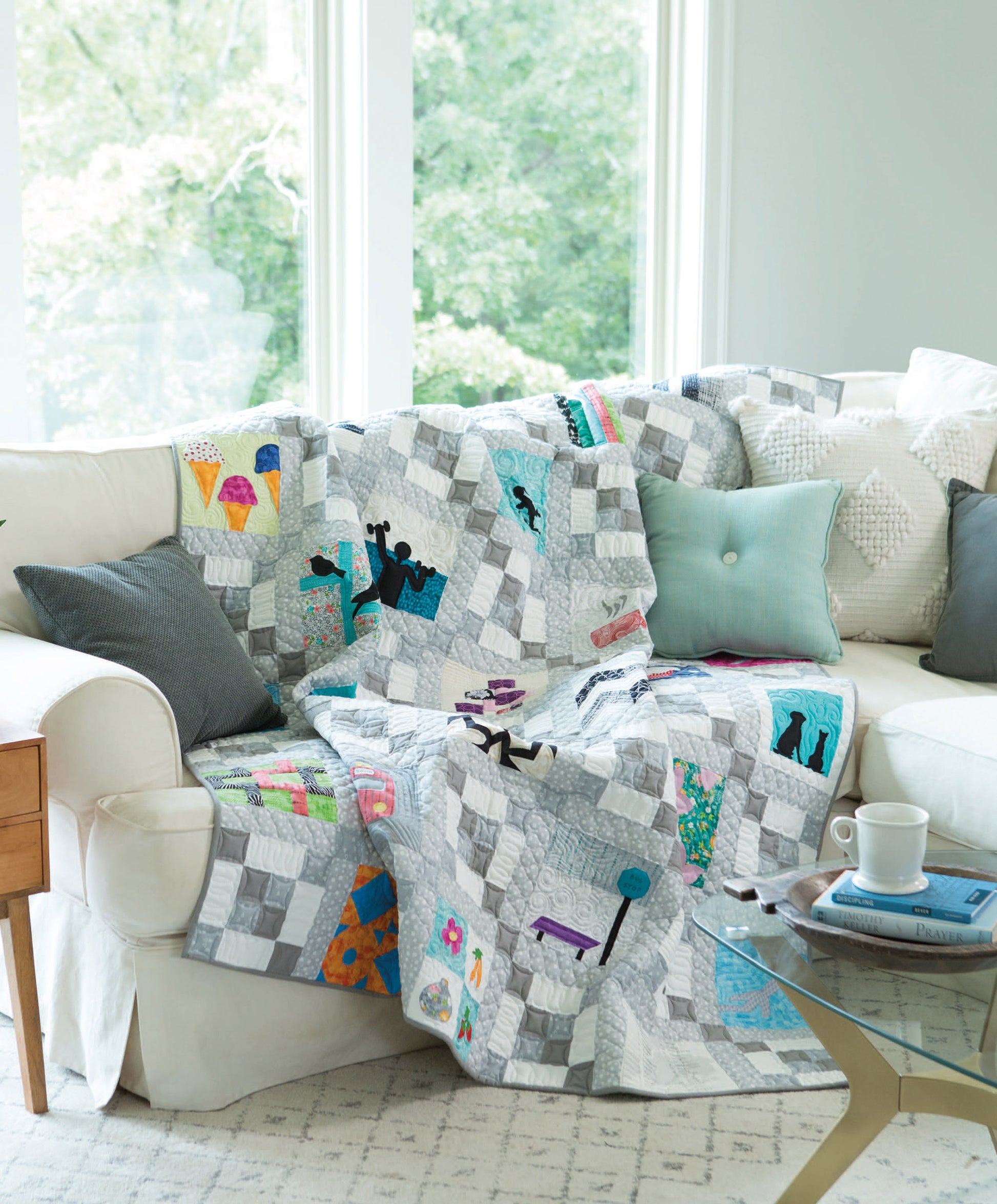 A vibrant City Blocks—32 Pieced and Applique Blocks Portraying City Life quilt from Leisure Arts is displayed over a white sectional with gray and aqua pillows in a sunlit living room, beside a side table with a coffee cup and book.