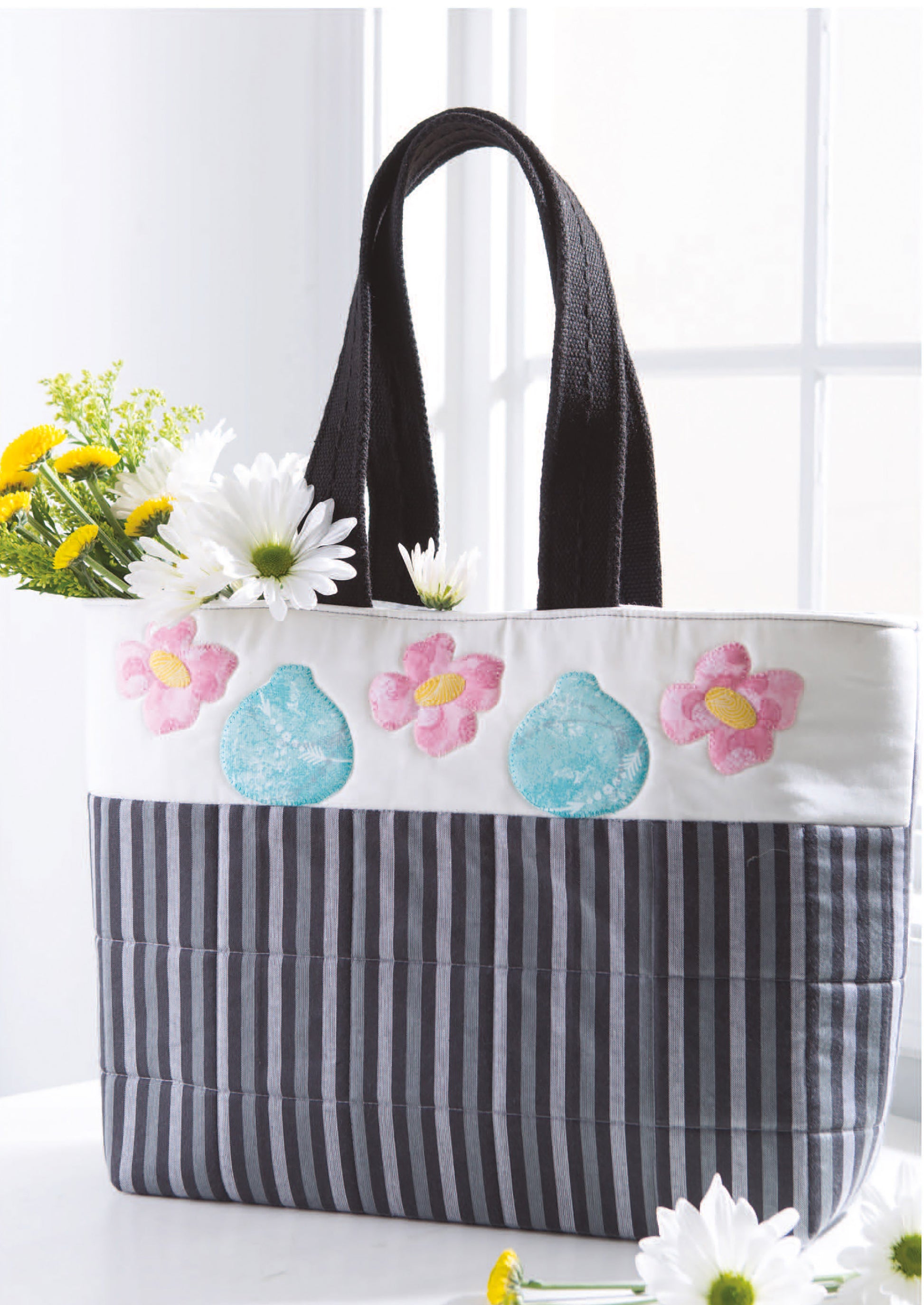 A cheerful tote decorated with blue vases and pink flowers, filled with daisies, sits by a bright window—perfect for carrying your City Blocks—32 Pieced and Applique Blocks Portraying City Life Digital Download from Leisure Arts.