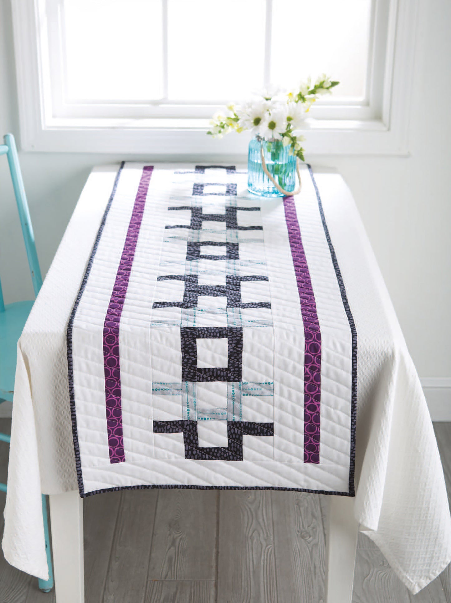 A white table with a modern quilted runner made using "City Blocks—32 Pieced and Applique Blocks Portraying City Life" by Leisure Arts features purple, blue, and black geometric patterns; a vase with white flowers and a turquoise chair are nearby.