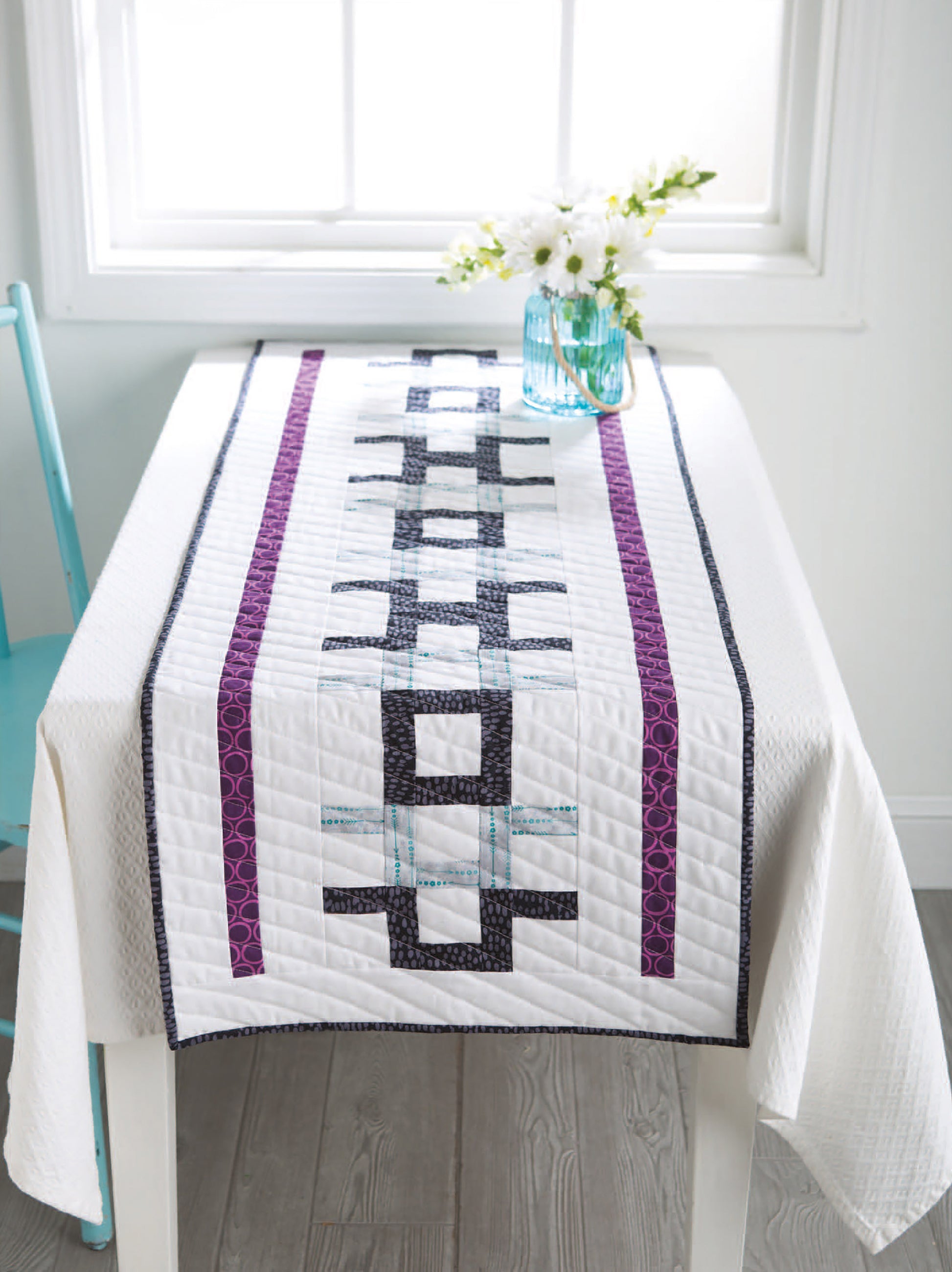 A white table with a modern quilted runner made using "City Blocks—32 Pieced and Applique Blocks Portraying City Life" by Leisure Arts features purple, blue, and black geometric patterns; a vase with white flowers and a turquoise chair are nearby.