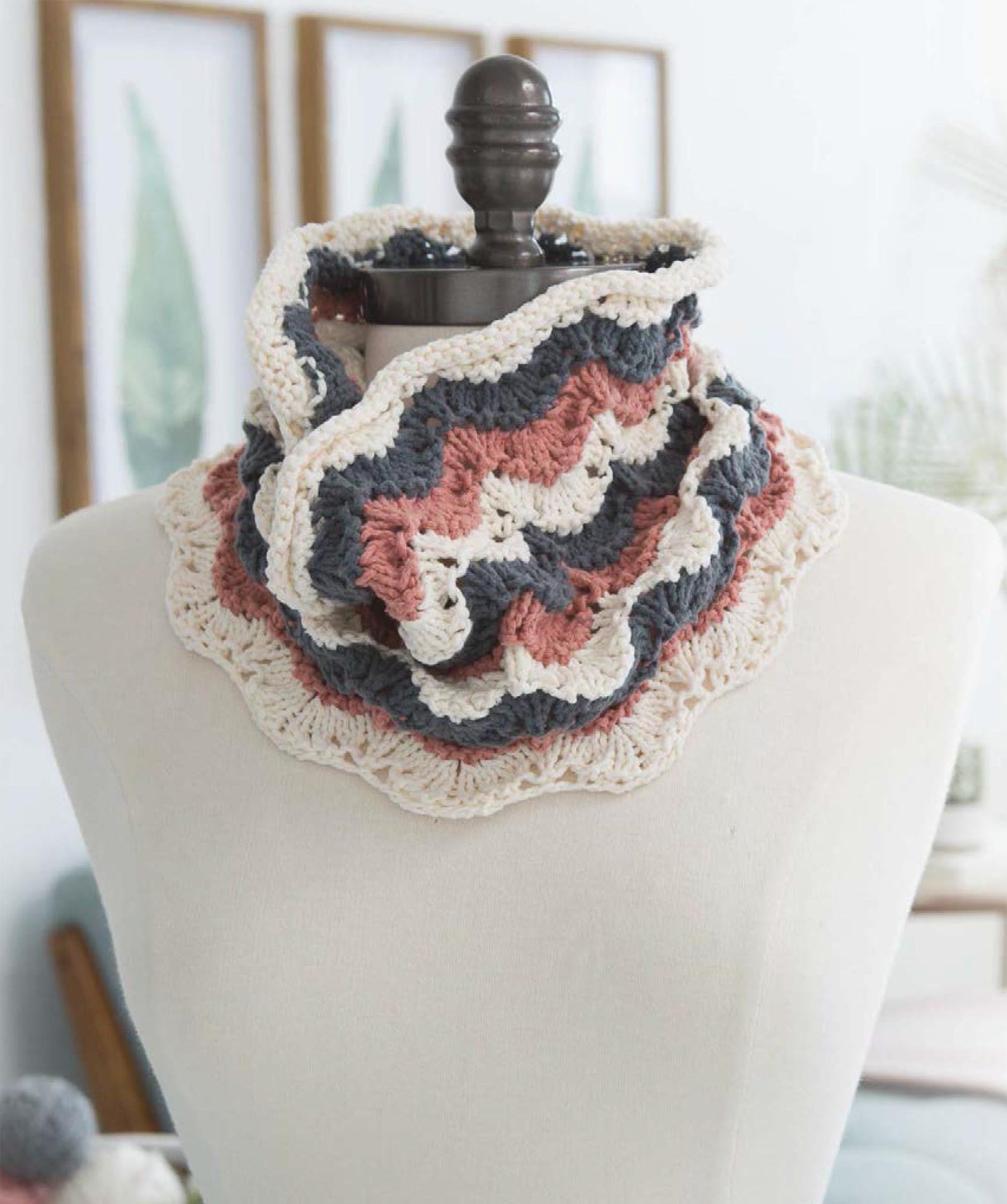 A mannequin displays the "Get Your Lace On" crocheted cowl by Leisure Arts, featuring wavy stripes in cream, dusty pink, and gray with a scalloped edge. Framed botanical prints and soft decor accent the background.