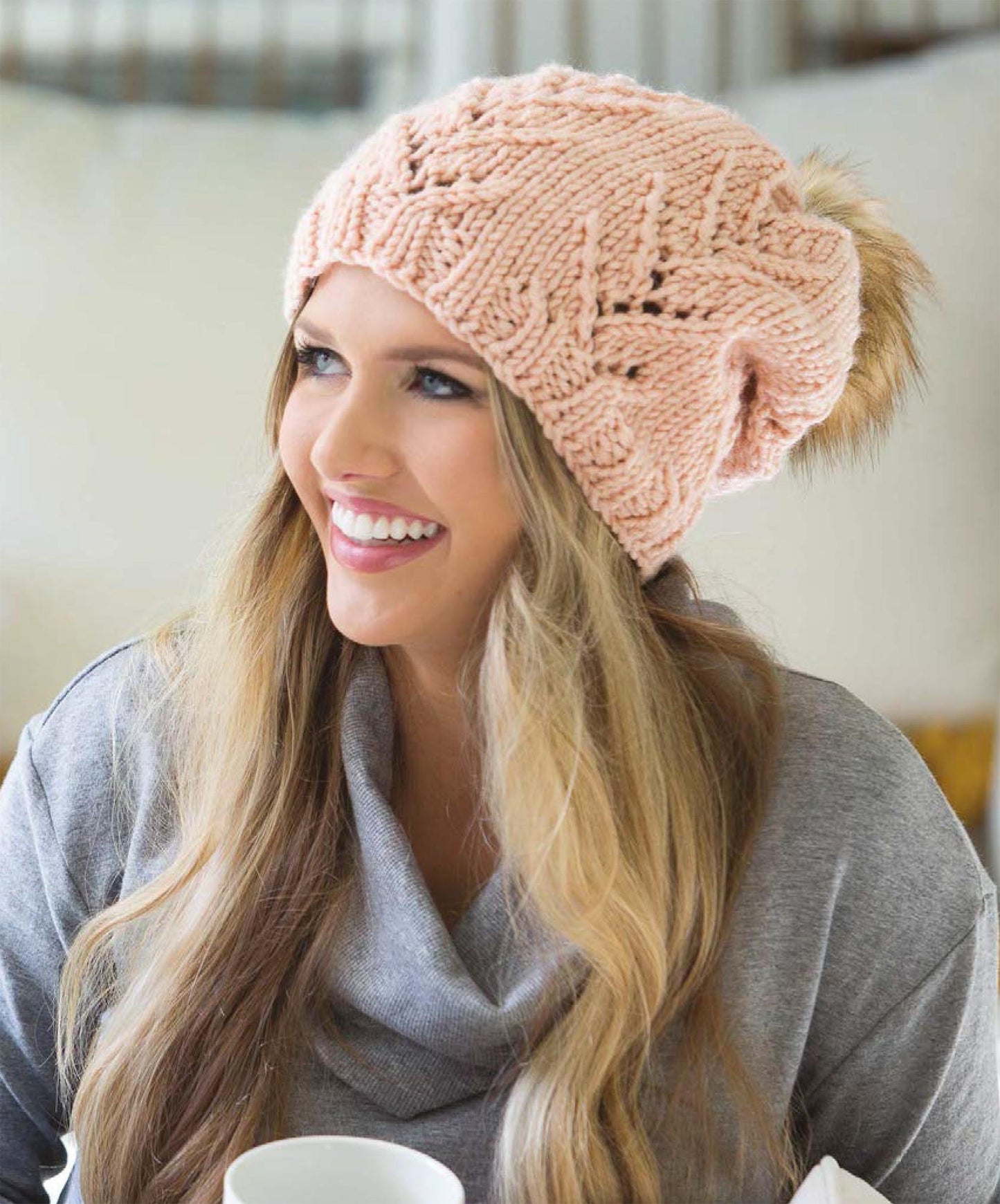 A smiling woman with long, wavy hair wears the 'Get Your Lace On' chunky light pink knitted beanie by Leisure Arts, featuring a fur pom-pom. She pairs it with a gray sweater, holds a white mug, and looks to the side.