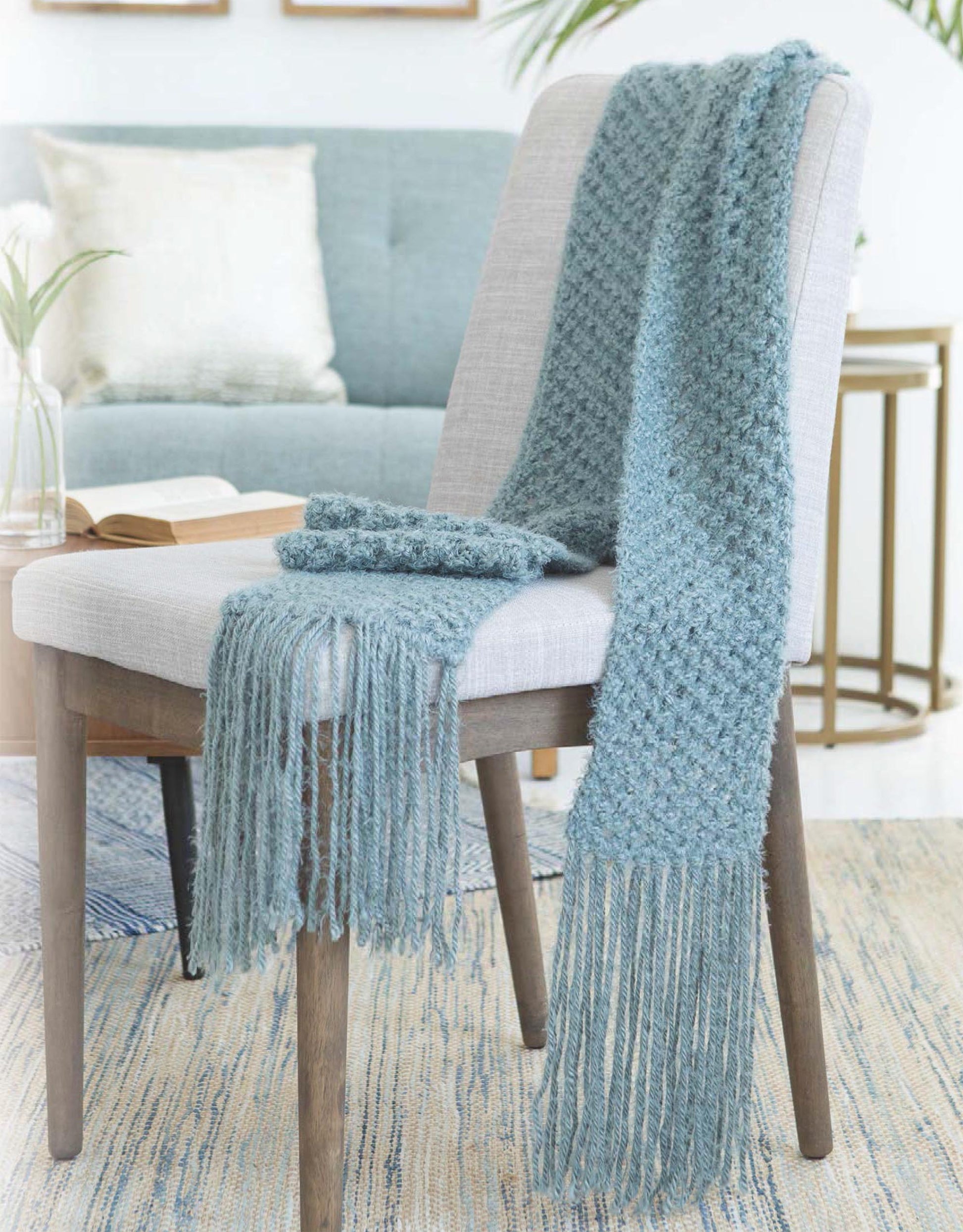 The Get Your Lace On scarf and matching mittens by Leisure Arts, in textured blue knit with long fringes, are displayed on a light gray chair—perfect for women's beginner knitting projects or cozy pattern explorations.