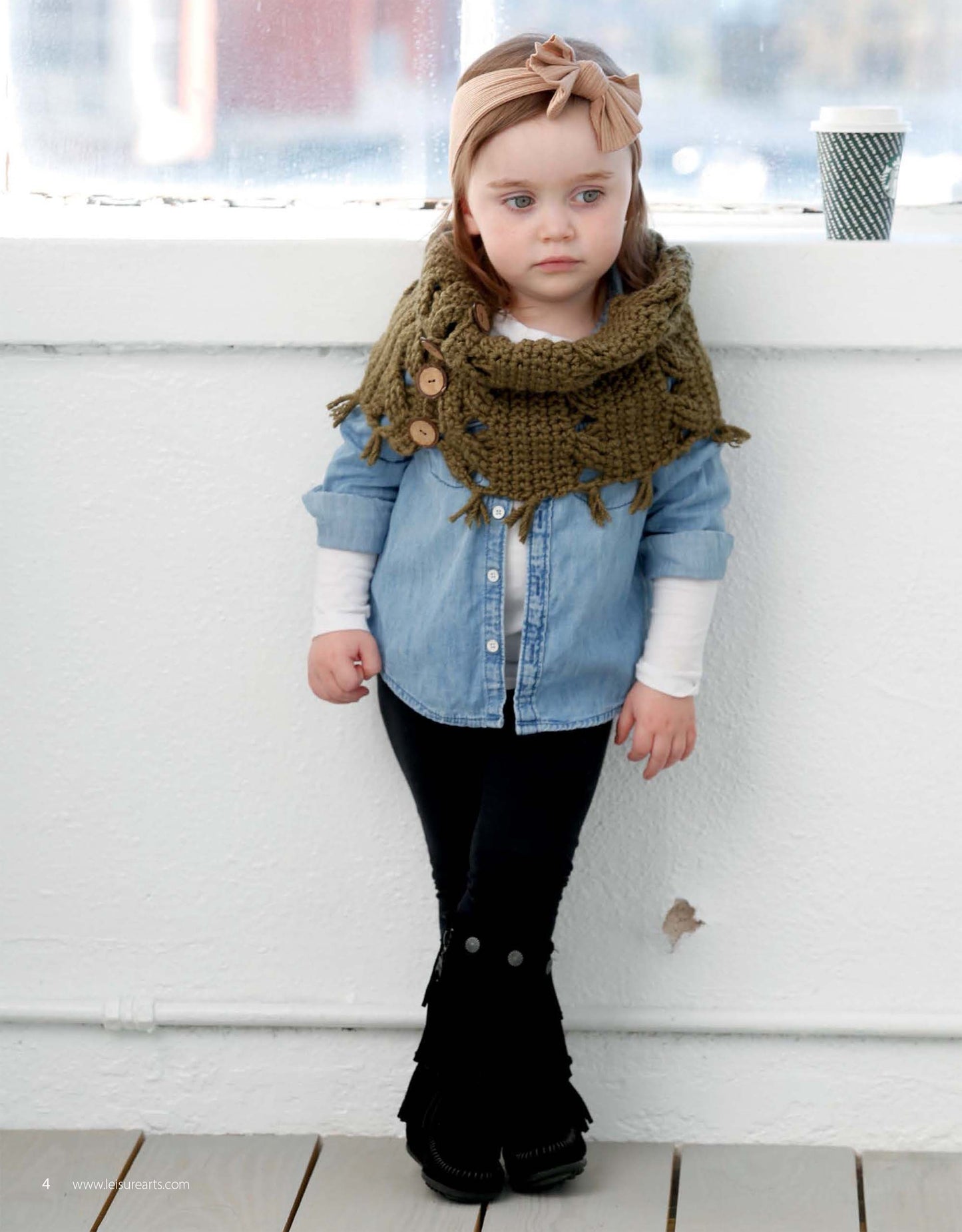 A young girl stands indoors against a white wall, modeling Leisure Arts’ Crochet Lil' Hipsters—an olive green buttoned scarf great for those seeking easy or beginner crochet patterns for toddlers.