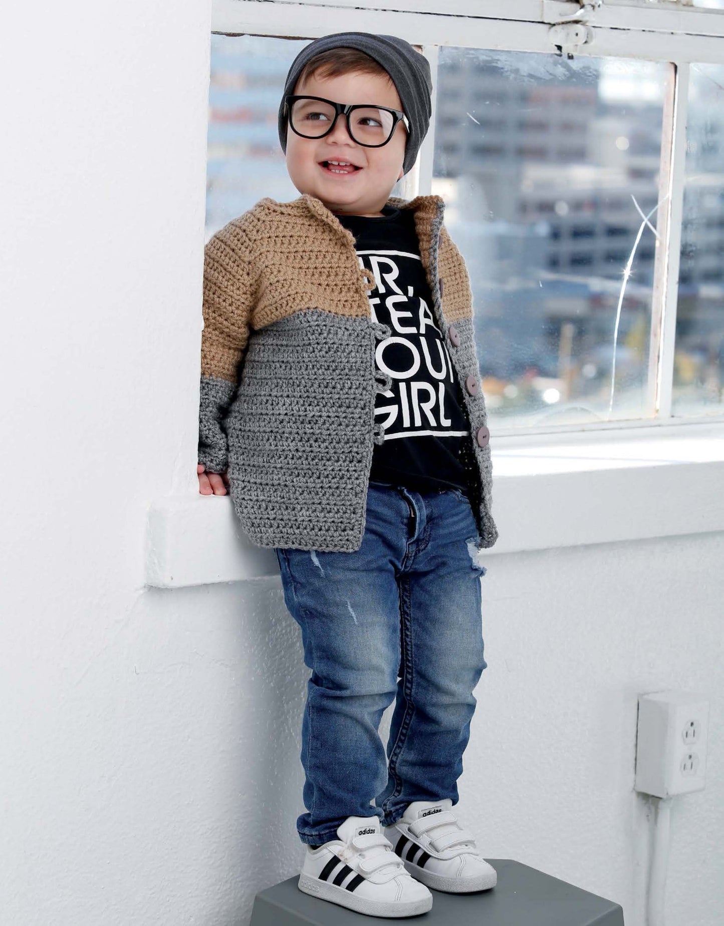 A smiling young child wearing glasses, a gray beanie, jeans, a black graphic T-shirt, white Adidas sneakers, and the Crochet Lil' Hipsters cardigan by Leisure Arts stands on a stool indoors by a window.