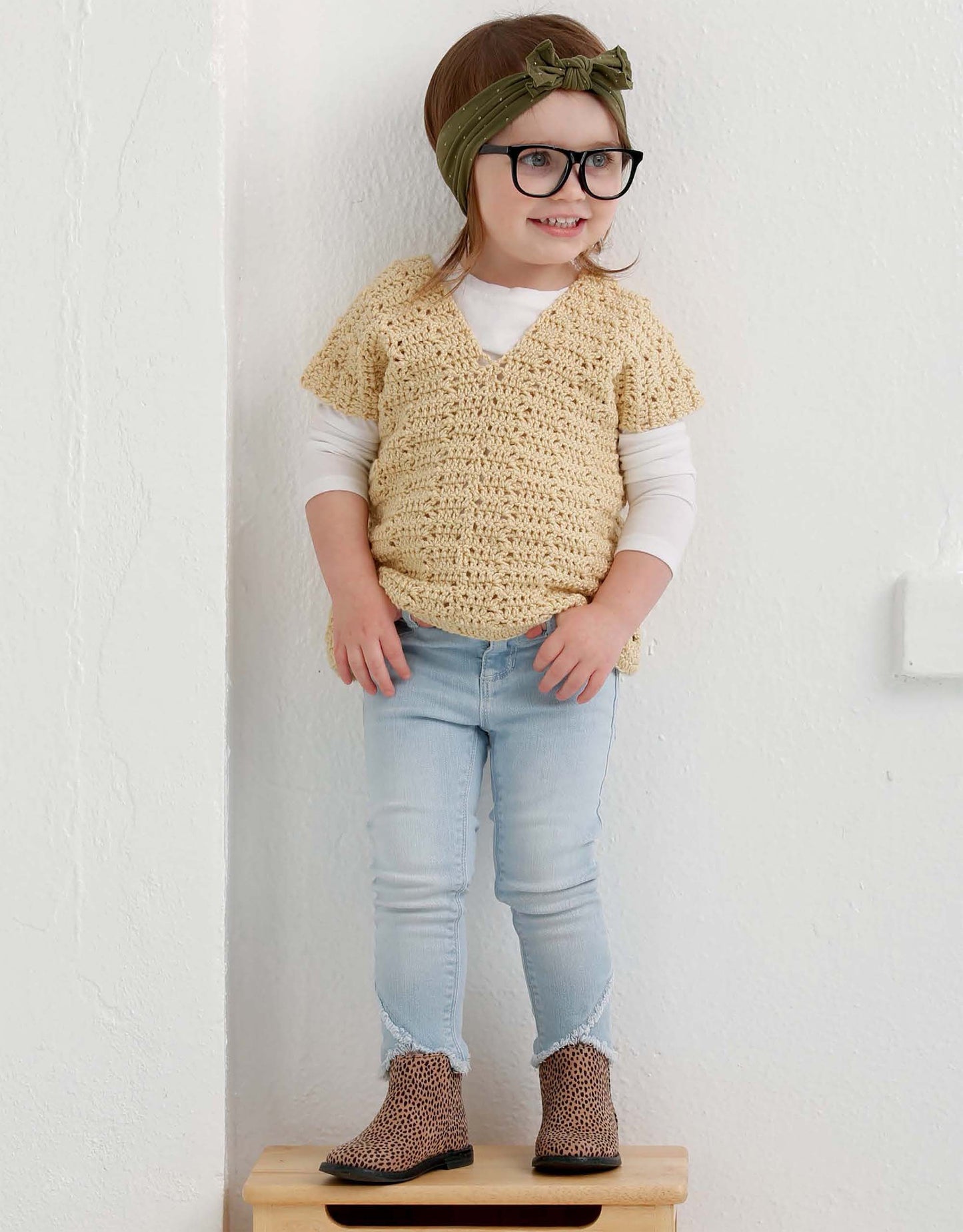 A young child, smiling against a plain white wall, stands on a small wooden stool wearing a beige sweater from the Leisure Arts Crochet Lil' Hipsters collection over a white shirt, jeans, leopard boots, large black glasses, and a green bow headband.