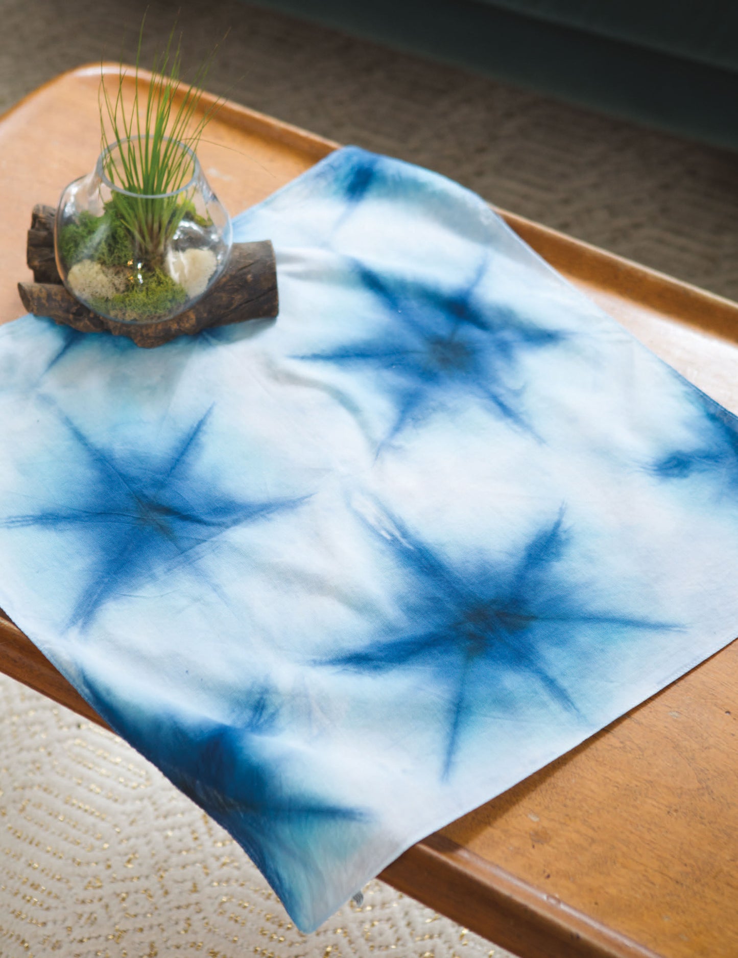 The "Shibori, Ice & Dip Dyeing" kit by Leisure Arts features a blue and white starburst-patterned cloth on a wooden table, with a glass bowl of plants, sand, and rocks displayed nearby atop driftwood. Perfect for beginner dyeing techniques.